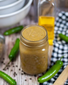 A jar of homemade green chili enchilada sauce on a wooden table, surrounded by fresh jalapeños, black pepper, and a bottle of oil.