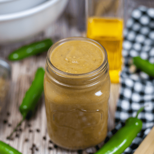 A jar of homemade green chili enchilada sauce on a wooden table, surrounded by fresh jalapeños, black pepper, and a bottle of oil.