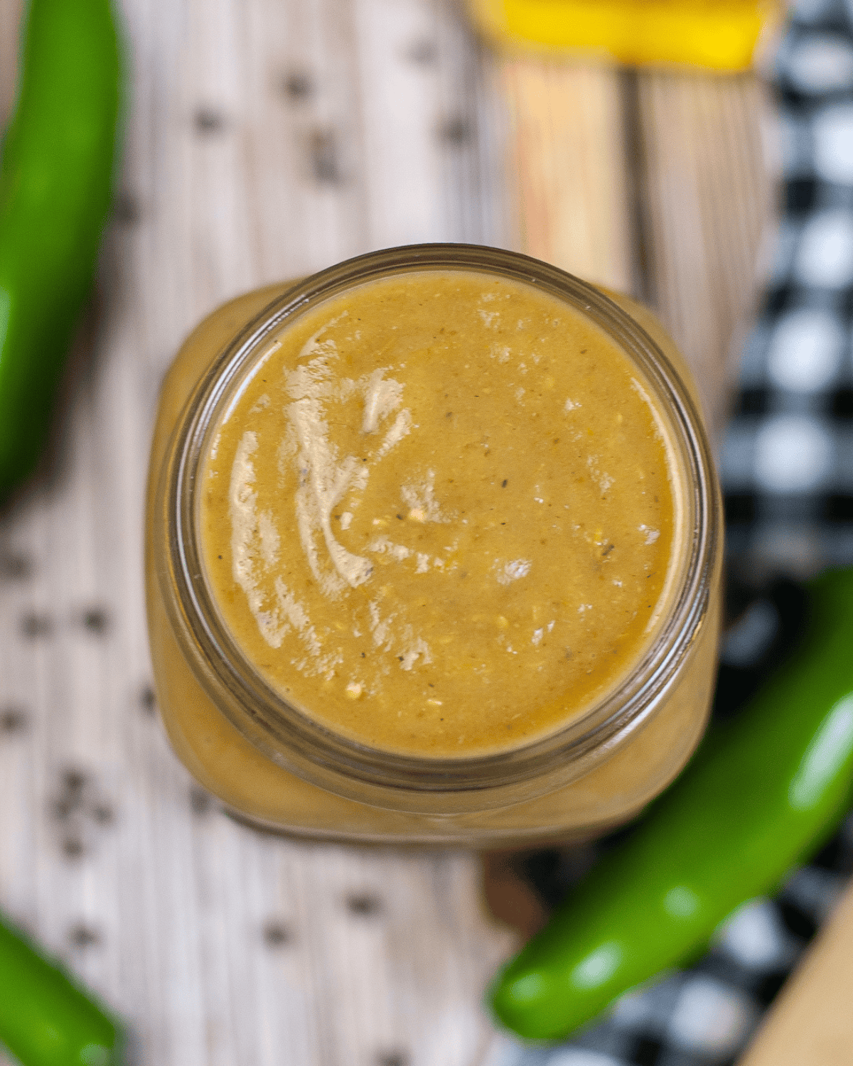 Overhead view of a jar of homemade green chili enchilada sauce surrounded by fresh jalapeno peppers and black-and-white checkered cloth.