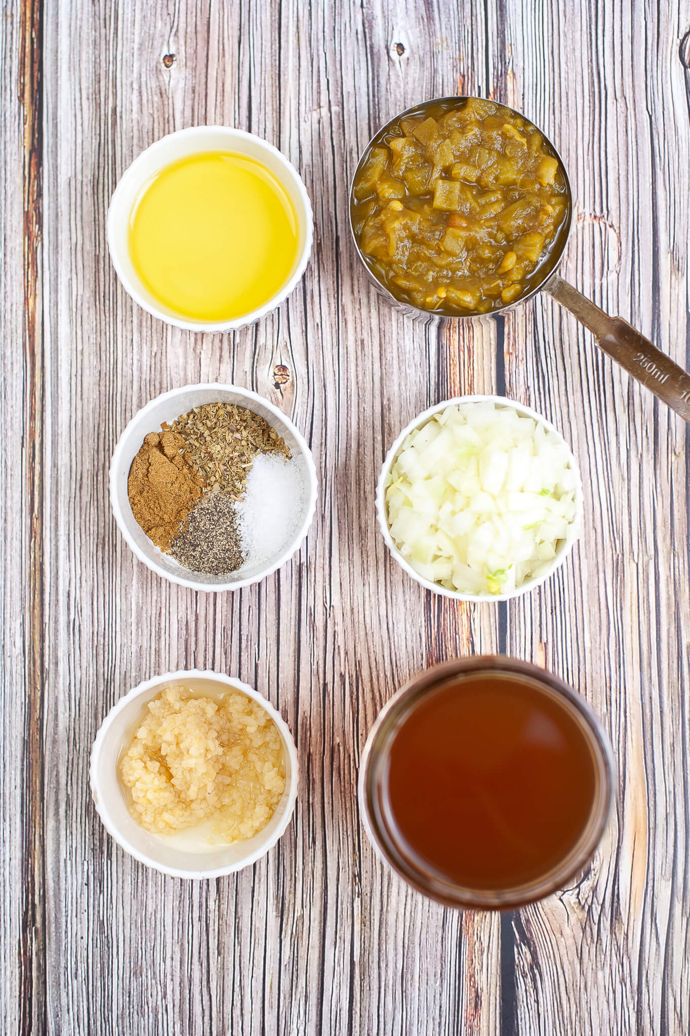Ingredients for cooking displayed on a wooden surface including bowls of oil, diced pickles, spices, chopped onions, and a jar of green chili enchilada sauce.