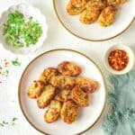 Two plates of oval-shaped homemade tater tots on a table, surrounded by small bowls containing chopped herbs, pepper, red pepper flakes, and a bowl of red sauce.