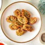 A white plate with several pieces of golden-brown Homemade Tater Tots arranged in a circle, placed on a light-colored surface with herbs, a green cloth, and a small bowl of pepper nearby.