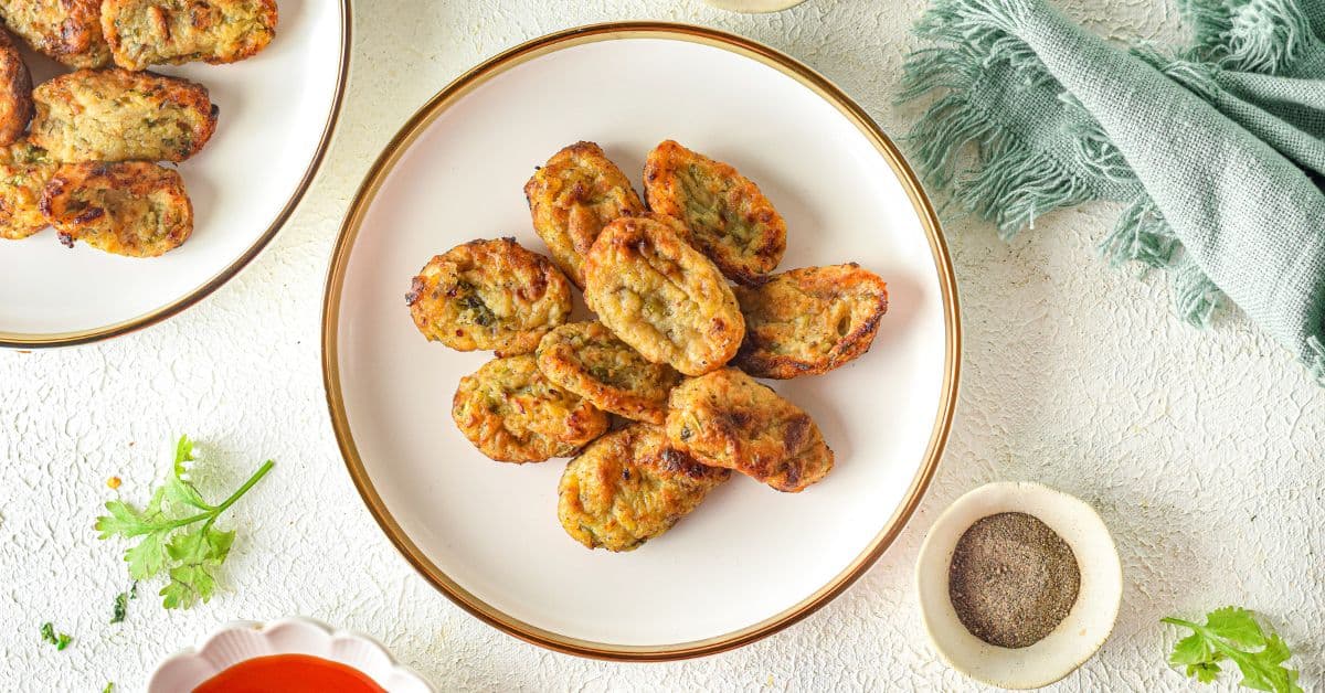 A white plate with several pieces of golden-brown Homemade Tater Tots arranged in a circle, placed on a light-colored surface with herbs, a green cloth, and a small bowl of pepper nearby.