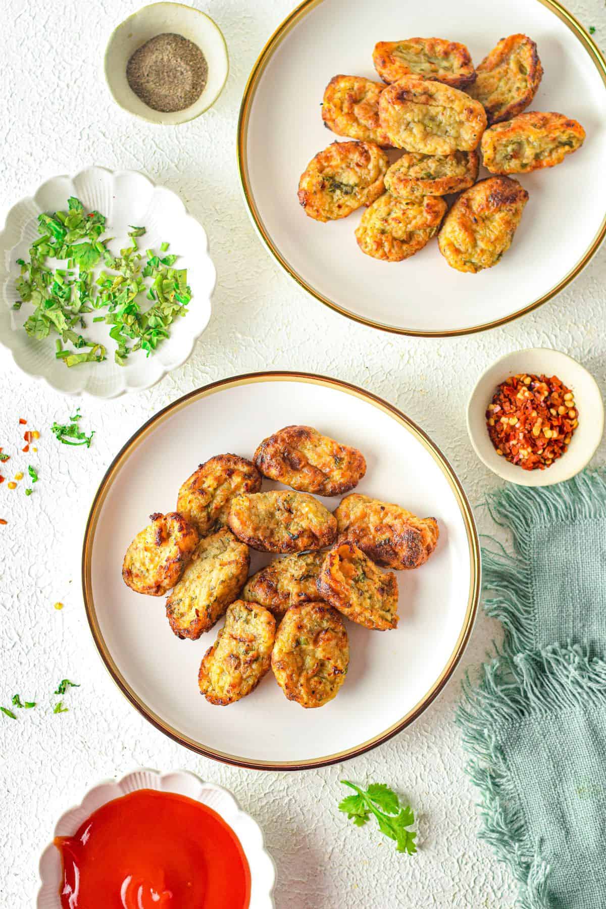 Two plates with oval-shaped potato rounds surrounded by small bowls of herbs, red chili flakes, black pepper, and ketchup on a white textured surface.