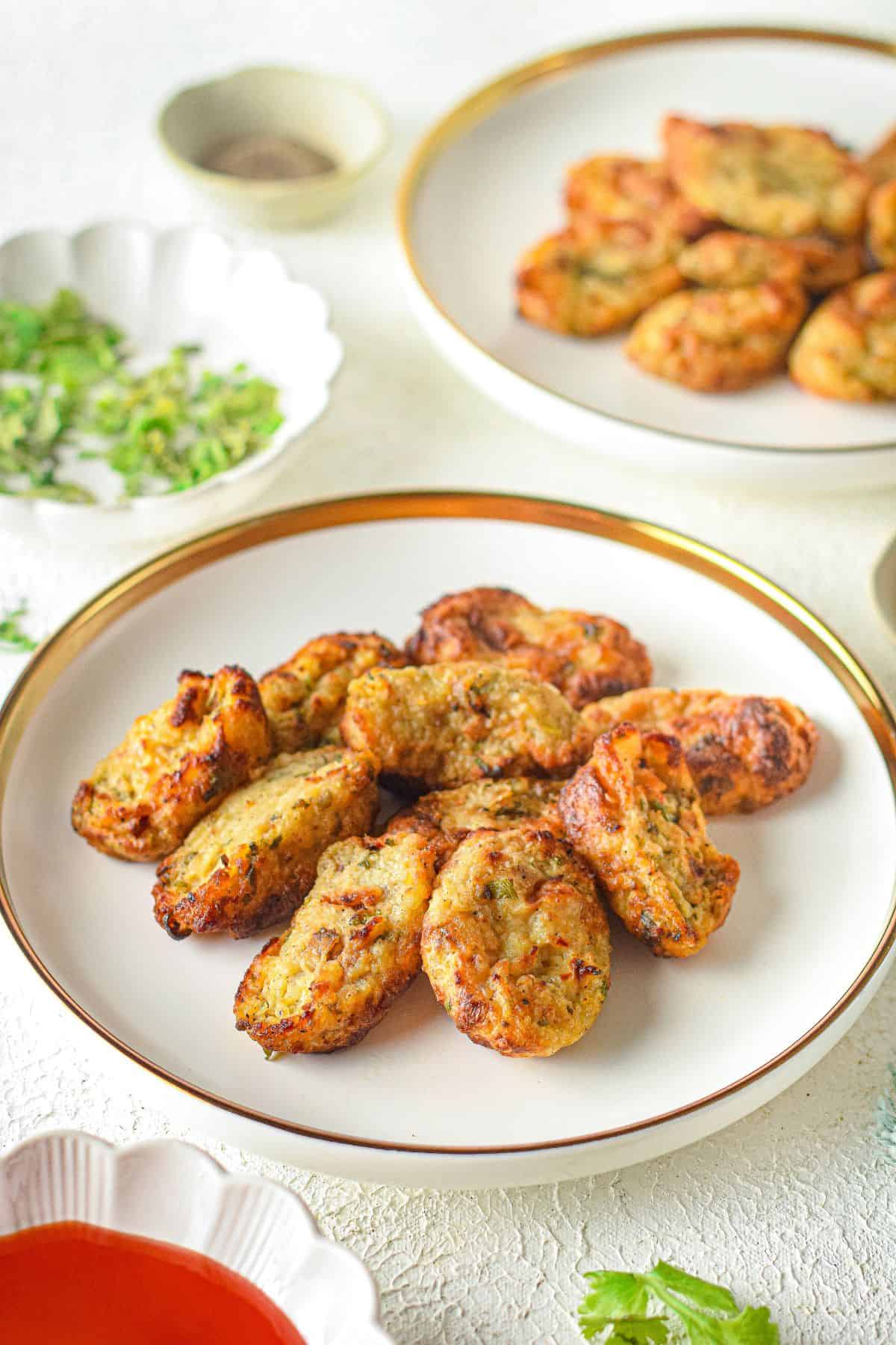 A white plate holds several golden-brown homemade tater tots; more fritters, chopped herbs, and dipping sauce are visible in the background.