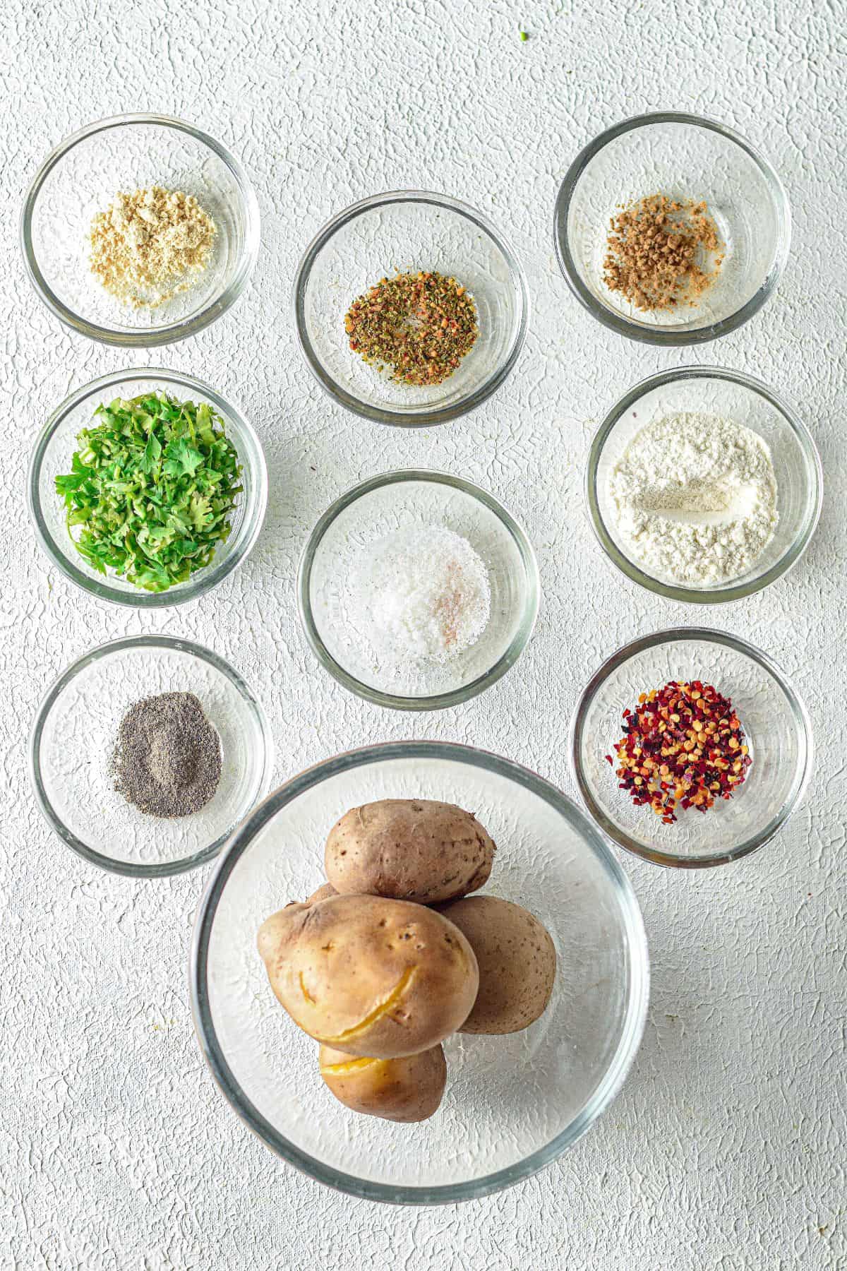 Nine glass bowls containing ingredients including whole potatoes, chopped herbs, flour, salt, pepper, chili flakes, and a variety of spices, all neatly arranged on a white surface.