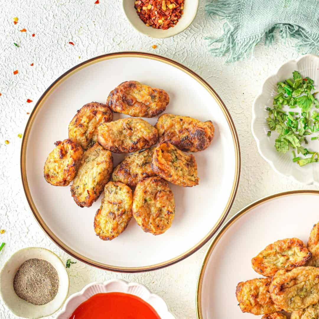 A plate of golden-brown homemade tater tots is surrounded by bowls of chopped herbs, red chili flakes, black pepper, and a dish of red sauce on a textured white surface.