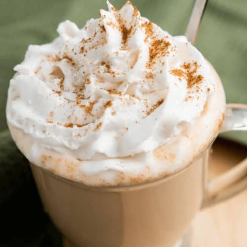A beige mug filled with a whipped cream-topped Iced Pumpkin Spice Latte, sprinkled with cinnamon, and a spoon on the right side. The mug is set on a light wooden surface with a green cloth partially visible.