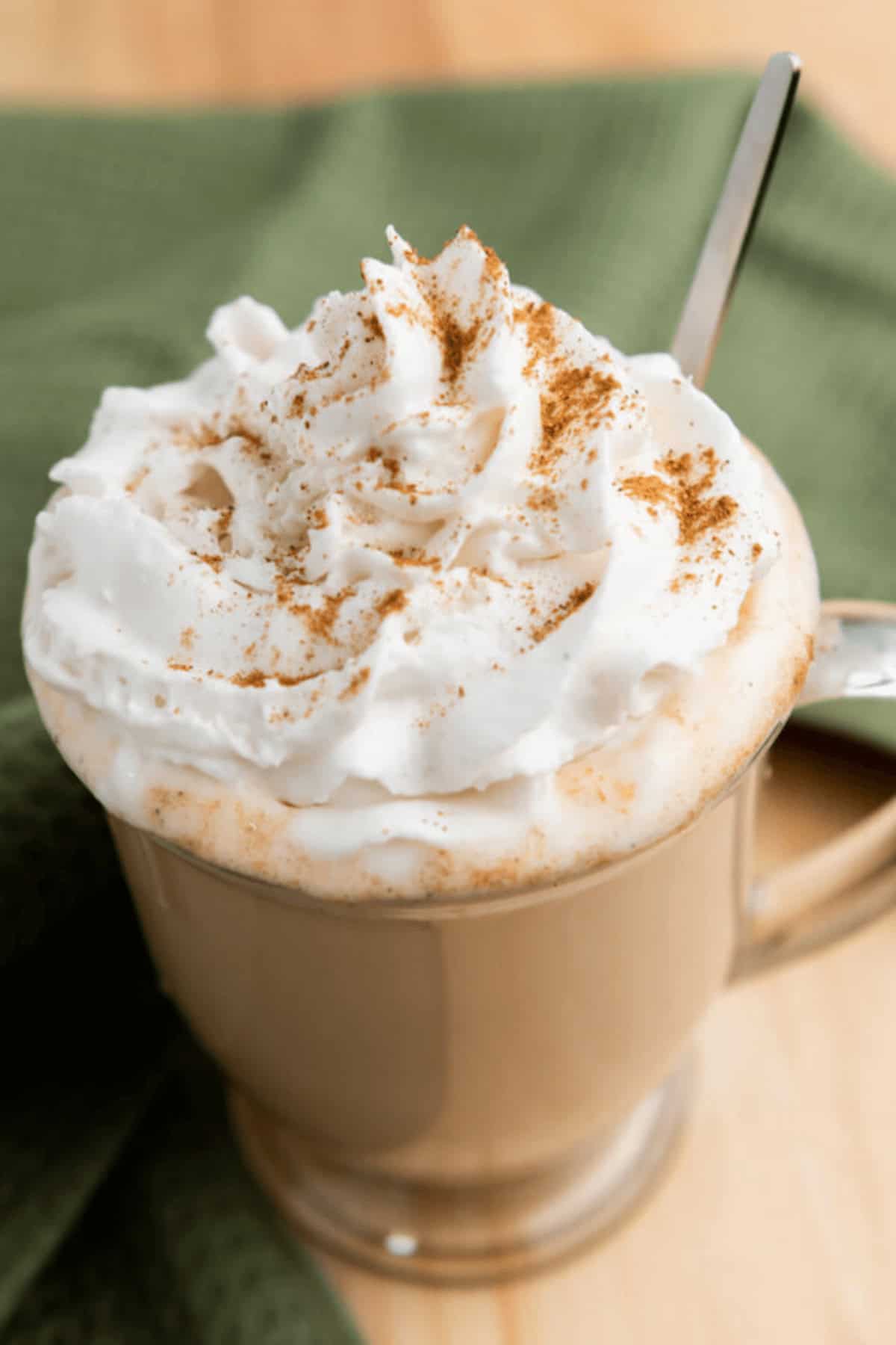 A beige mug filled with a whipped cream-topped Iced Pumpkin Spice Latte, sprinkled with cinnamon, and a spoon on the right side. The mug is set on a light wooden surface with a green cloth partially visible.