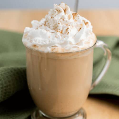 A glass mug filled with a frothy iced pumpkin spice latte topped with whipped cream and a sprinkle of cinnamon, placed on a wooden table with a green cloth nearby.