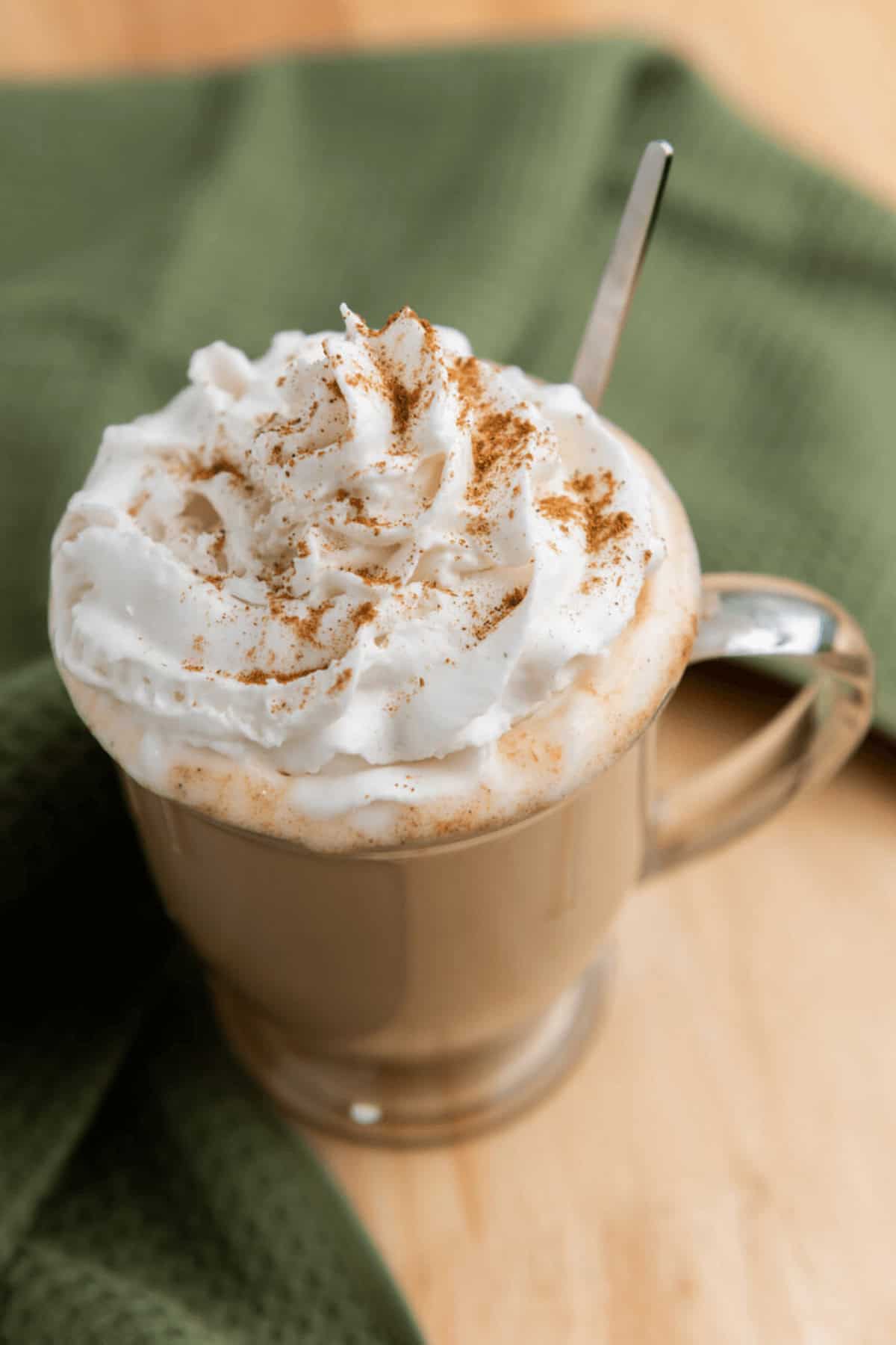 A glass mug filled with a whipped cream-topped Iced Pumpkin Spice Latte, sprinkled with cinnamon, placed on a wooden surface next to a green cloth.