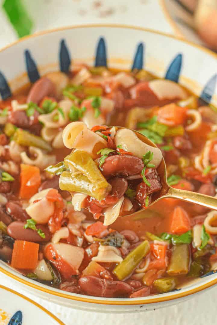 A bowl of Italian minestrone soup brimming with pasta, kidney beans, carrots, and green beans. A spoon is lifting a delicious portion of the classic soup.