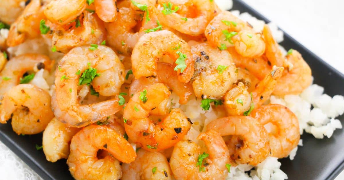 Roasted shrimp garnished with chopped parsley served on a bed of white rice on a black rectangular plate.