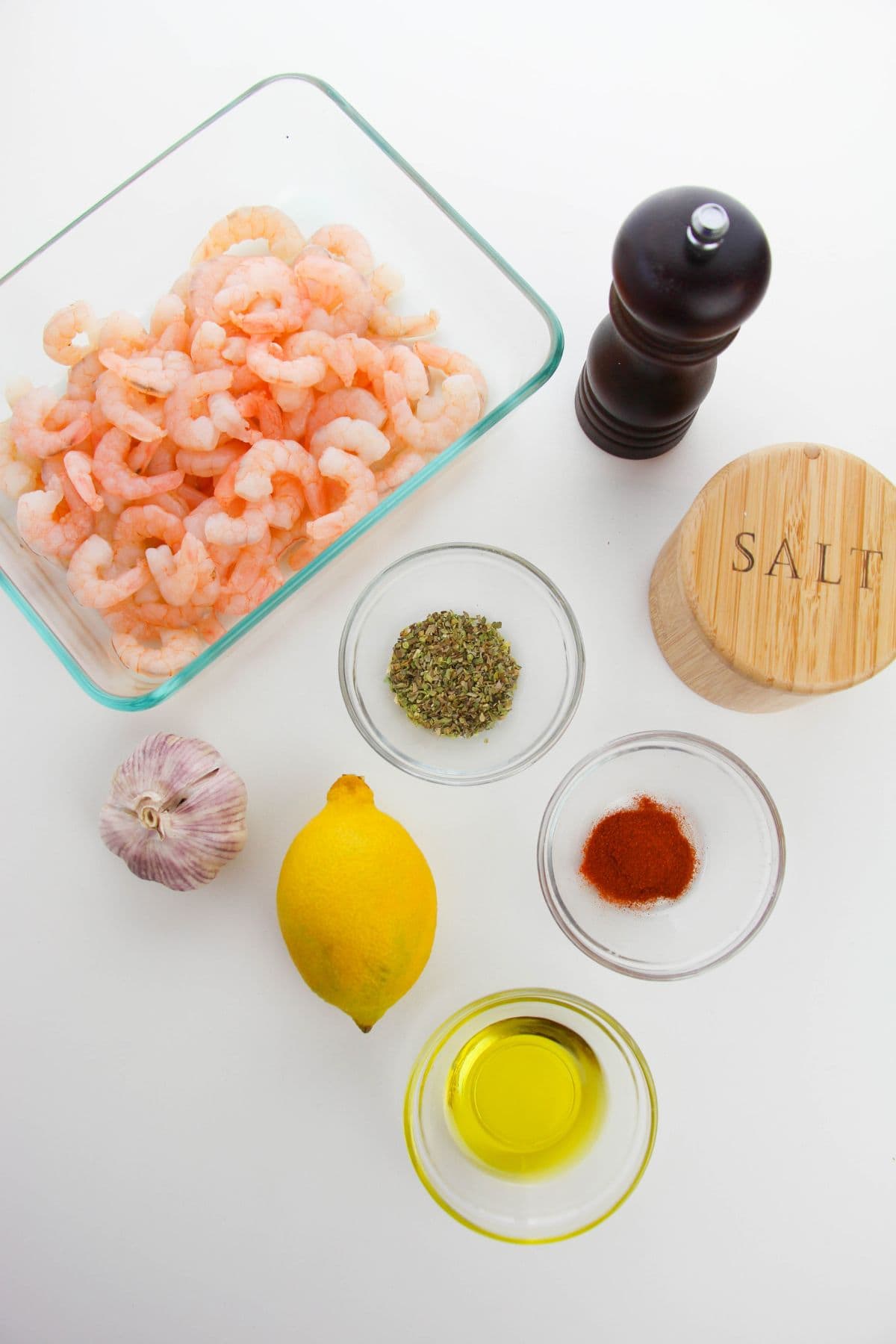 A glass container of seafood, a pepper grinder, salt jar, garlic bulb, lemon, olive oil, dried herbs, and paprika in small bowls on a white surface.