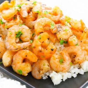 Roasted shrimp served on a bed of rice, garnished with chopped parsley on a black rectangular plate.