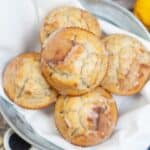 A bowl lined with a white napkin holds six Lemon Poppy Seed Cupcakes, with whole lemons and a blue checkered cloth beside it.