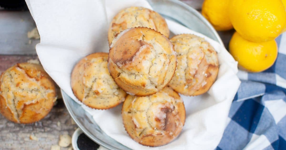 A bowl lined with a white napkin holds six Lemon Poppy Seed Cupcakes, with whole lemons and a blue checkered cloth beside it.