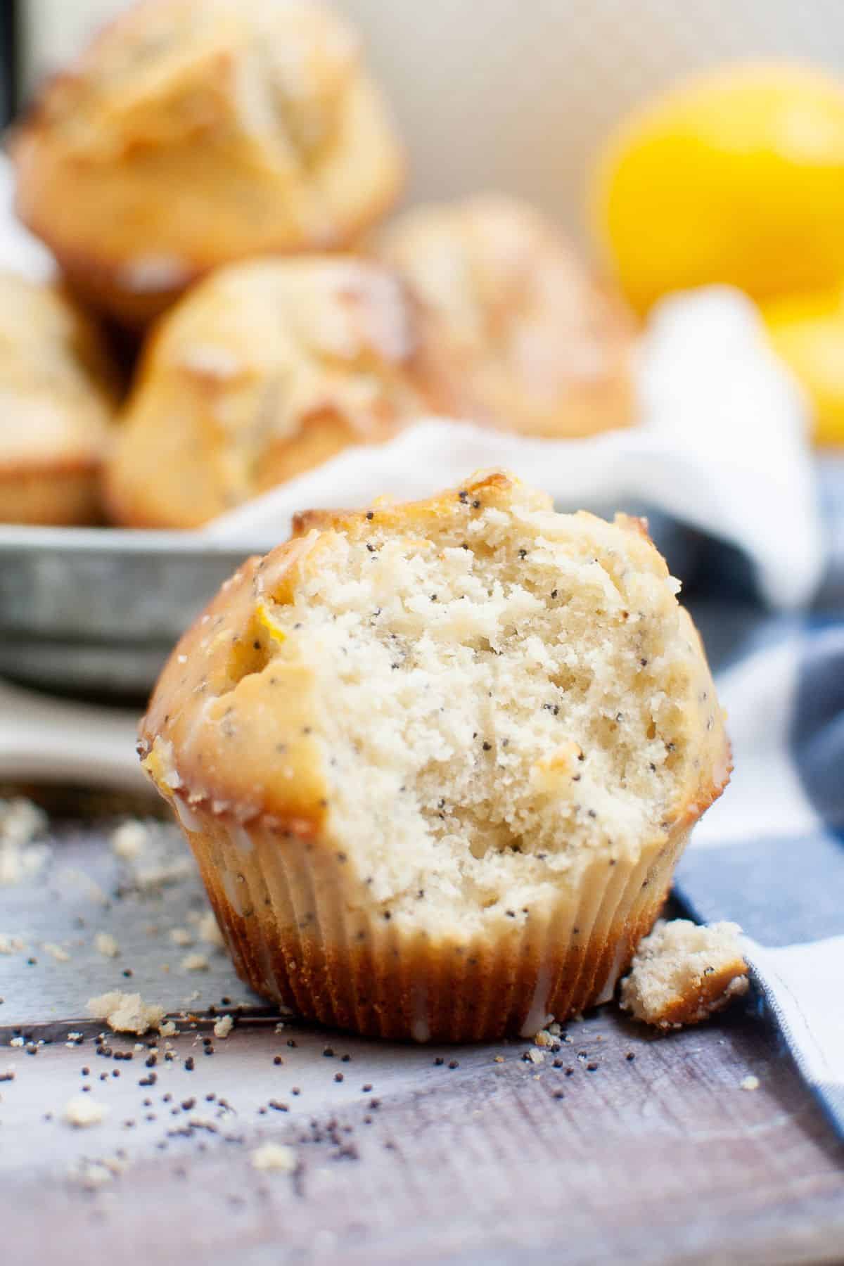 A close-up of a partially eaten muffin on a wooden surface, with scattered crumbs and whole muffins softly blurred in the background.