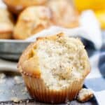 A partially eaten Lemon Poppy Seed Cupcake with visible poppy seeds sits on a wooden surface, with more cupcakes and a lemon in the background.