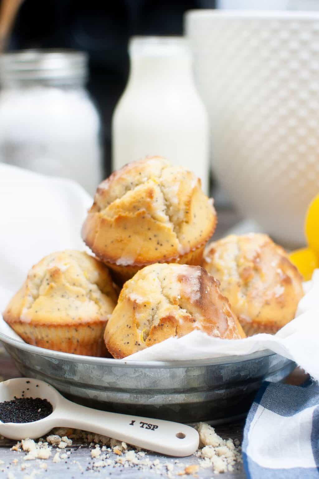 A metal bowl lined with a white cloth holds five glazed Lemon Poppy Seed Cupcakes. A measuring spoon with poppy seeds and baking ingredients are visible in the background.