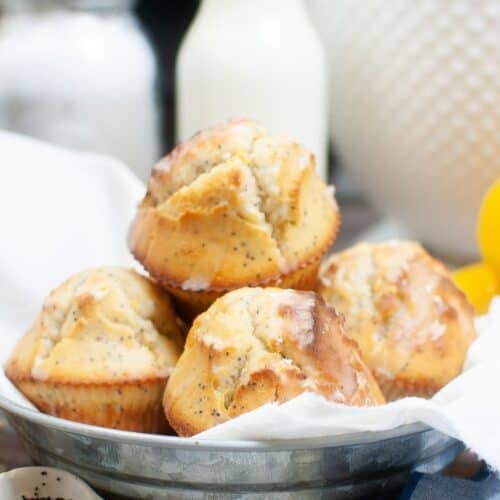 A metal bowl lined with a white cloth holds five glazed Lemon Poppy Seed Cupcakes. A measuring spoon with poppy seeds and baking ingredients are visible in the background.