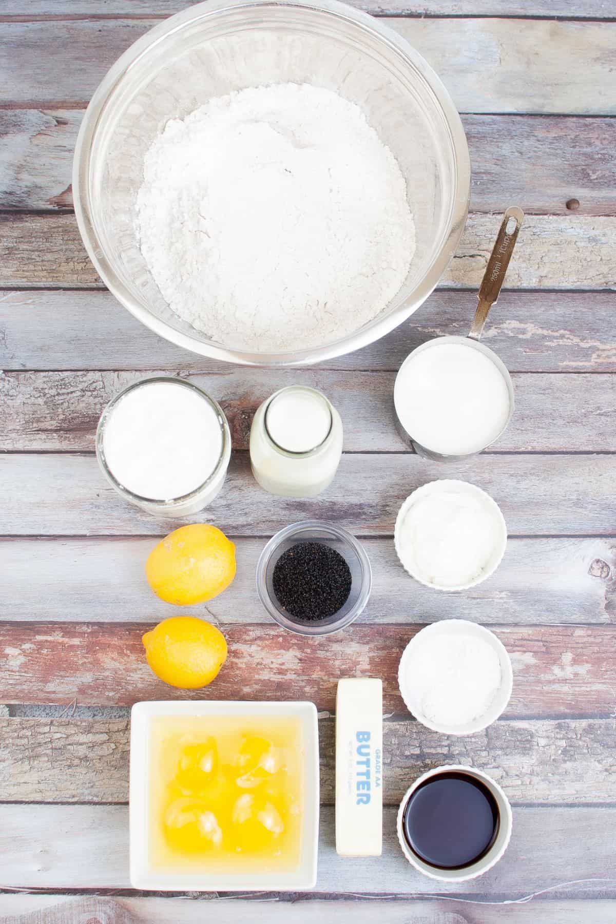 Overhead view of baking ingredients  including flour, sugar, eggs, butter, lemons, poppy seeds, milk, yogurt, and vanilla extract neatly arranged on a wooden surface.
