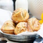 A metal bowl filled with four glazed Lemon Poppy Seed Cupcakes, accompanied by a jug of milk and a white cloth in the background.
