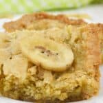A close-up of a Peanut Butter Banana Bar with a banana slice on top, displayed on a white plate with a green checkered cloth in the background.