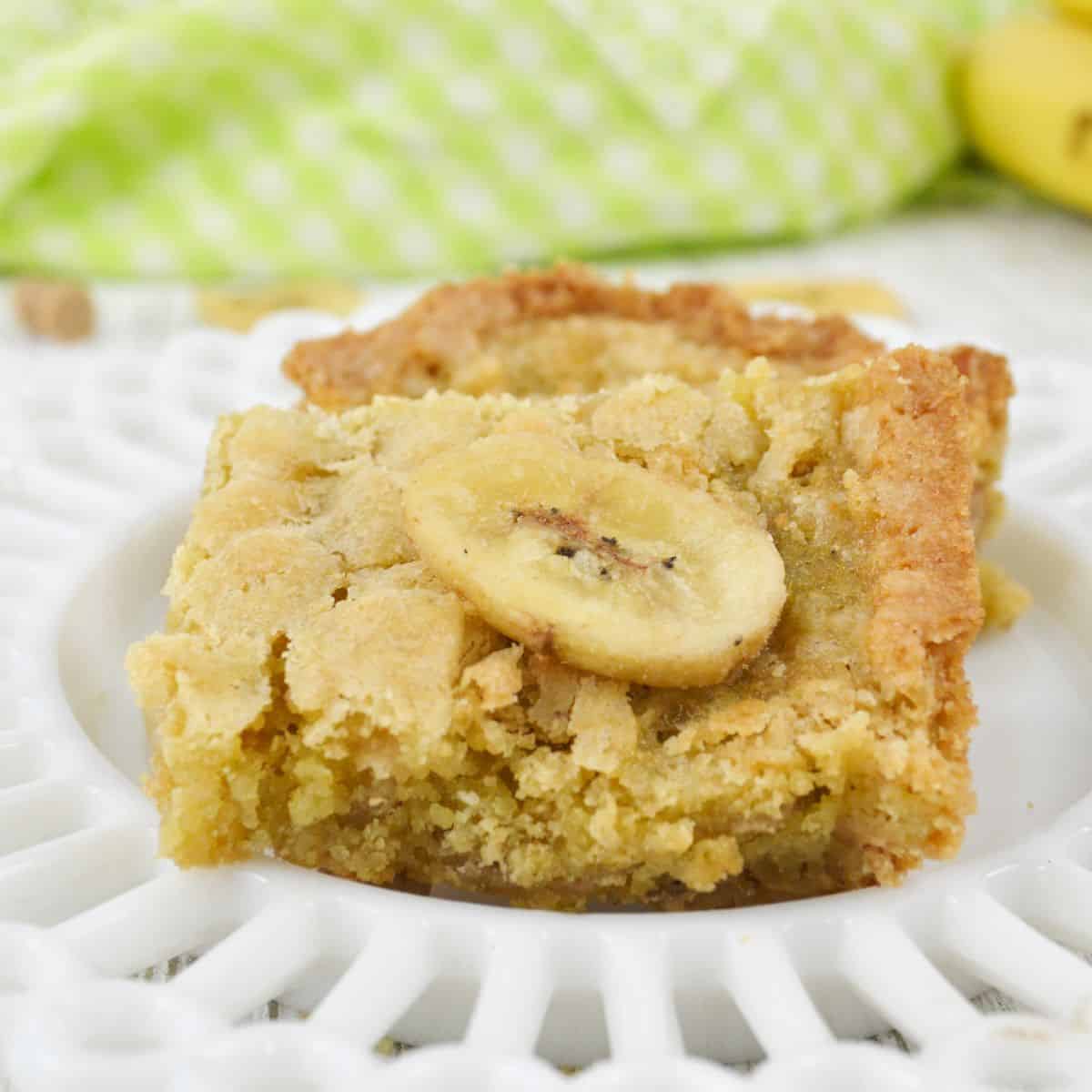 A close-up of a Peanut Butter Banana Bar topped with a banana chip, served on a white decorative plate.