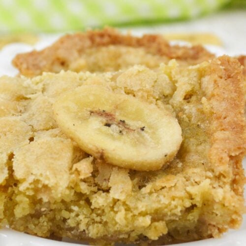 A close-up of a square Peanut Butter Banana Bar topped with a banana slice, displayed on a white plate with a green checkered cloth in the background.
