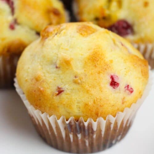 A close-up of a freshly baked Raspberry White Chocolate Muffin with visible pieces of fruit, wrapped in a white paper liner, sitting on a white surface.