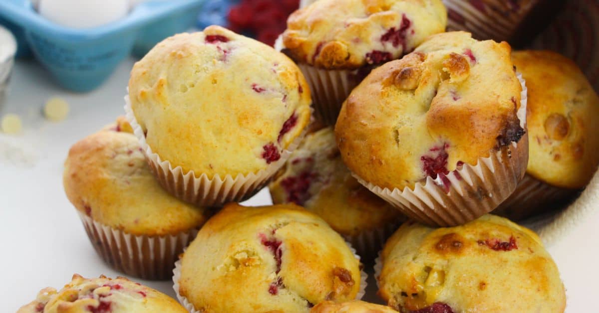 A close-up of Raspberry White Chocolate Muffins, showcasing a pile of golden-brown treats with visible pieces of raspberries and white chocolate in paper liners.