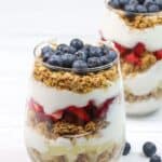 A glass filled with layers of granola, yogurt, sliced strawberries, and topped with fresh blueberries creates a vibrant Yogurt Parfait; another similar parfait sits in the background.