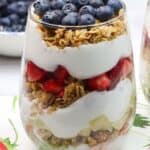 A glass filled with layers of granola, yogurt, diced strawberries, and topped with blueberries creates a delightful Yogurt Parfait, placed on a napkin with a strawberry print.