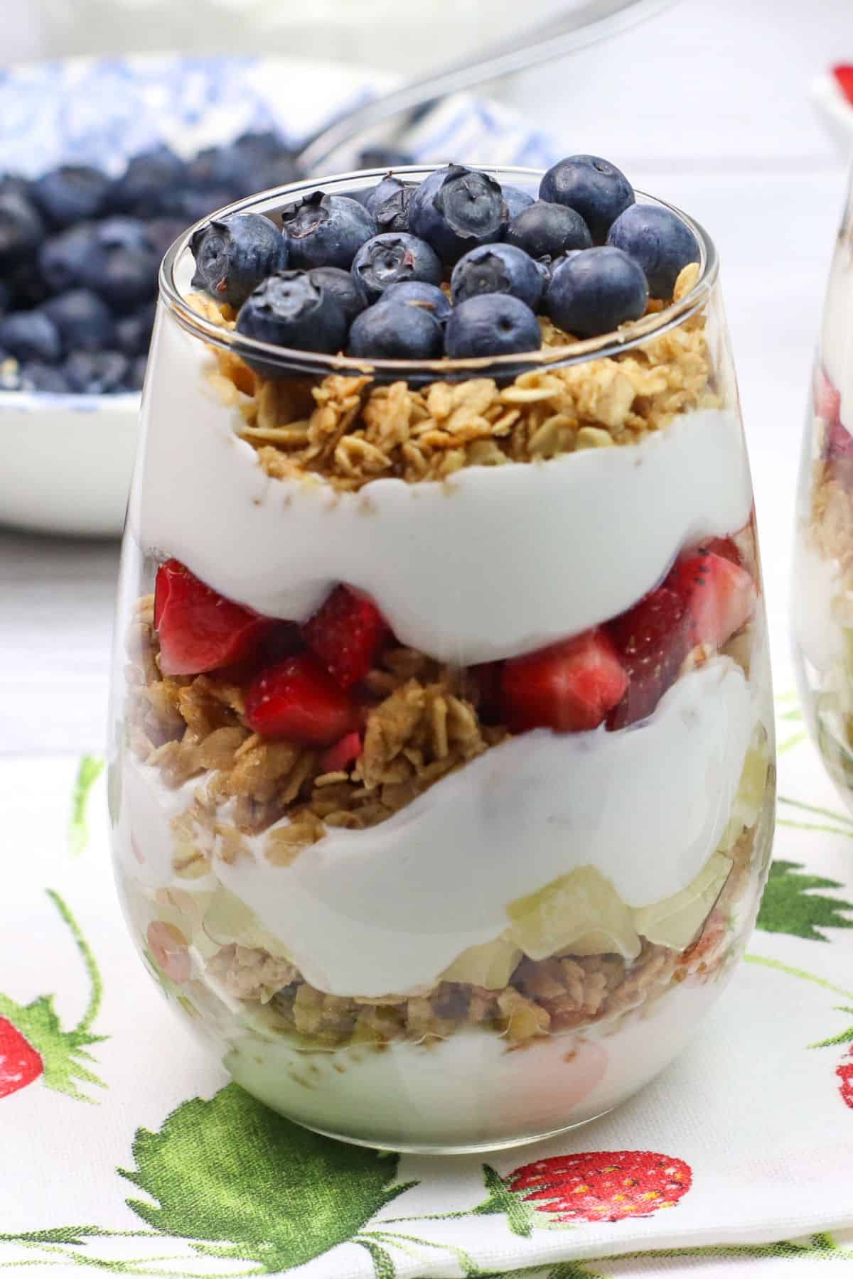 A glass filled with layers of granola, yogurt, diced strawberries, and topped with blueberries creates a delightful Yogurt Parfait, placed on a napkin with a strawberry print.