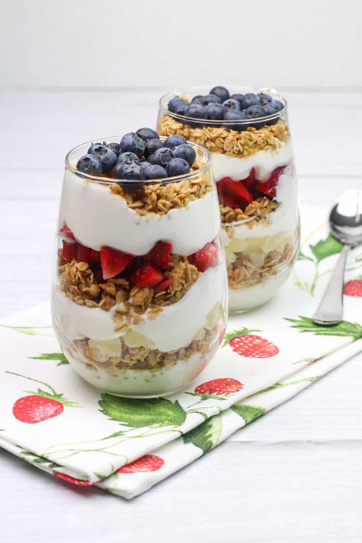 Two glass cups filled with layers of granola, creamy yogurt, strawberries, and blueberries sit on a strawberry-patterned napkin next to two spoons, creating an inviting Yogurt Parfait display.