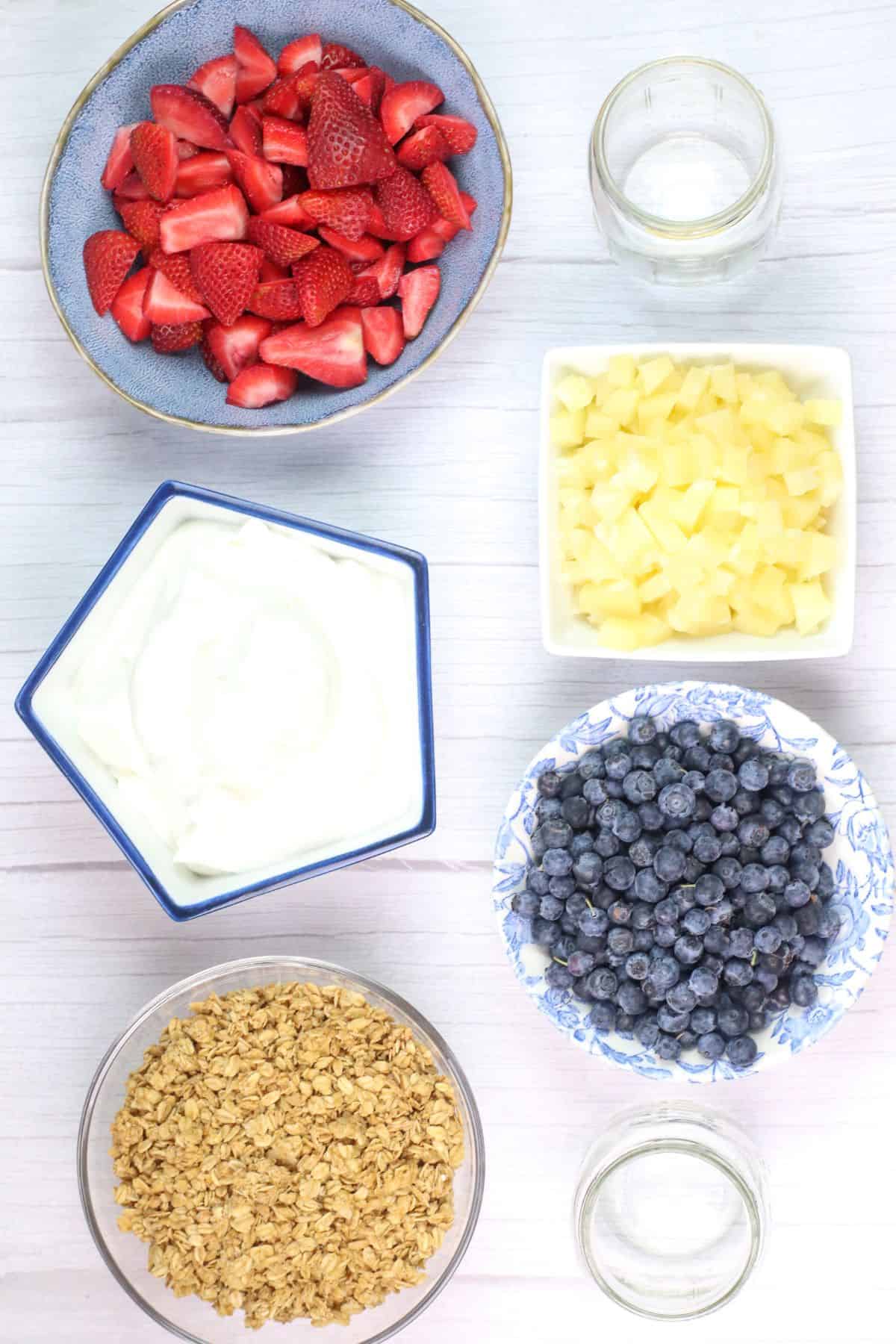 Bowls of sliced strawberries, pineapple chunks, blueberries, granola, and yogurt arranged on a white surface invite you to create your own Yogurt Parfait, with two empty glass jars ready for layering.