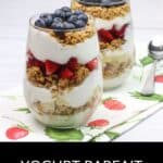 Two glasses of yogurt parfait, layered with creamy yogurt, granola, strawberries, and blueberries, sit on a strawberry-print napkin beside two spoons.
