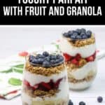 Two glasses of yogurt parfait are filled with layers of yogurt, granola, strawberries, bananas, and blueberries on a light surface, with extra blueberries scattered nearby.