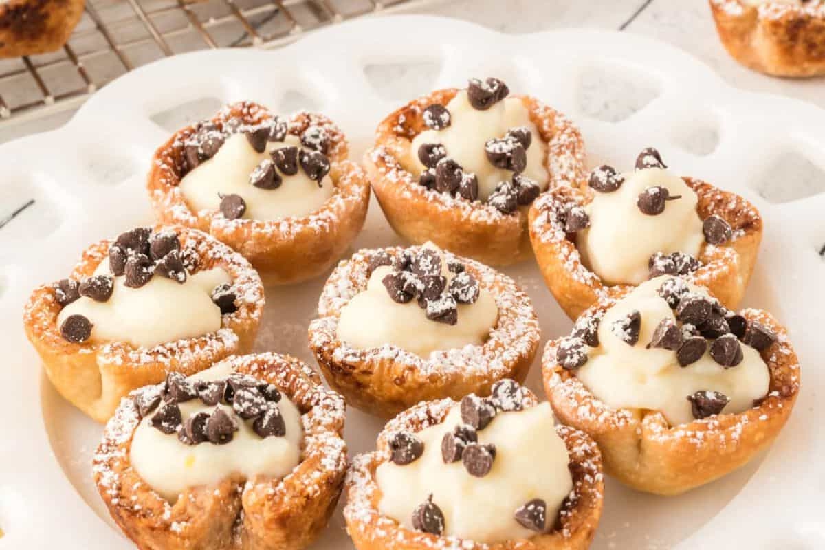  Mini cannoli cups filled with creamy ricotta filling, topped with mini chocolate chips and dusted with powdered sugar, arranged on a white plate.