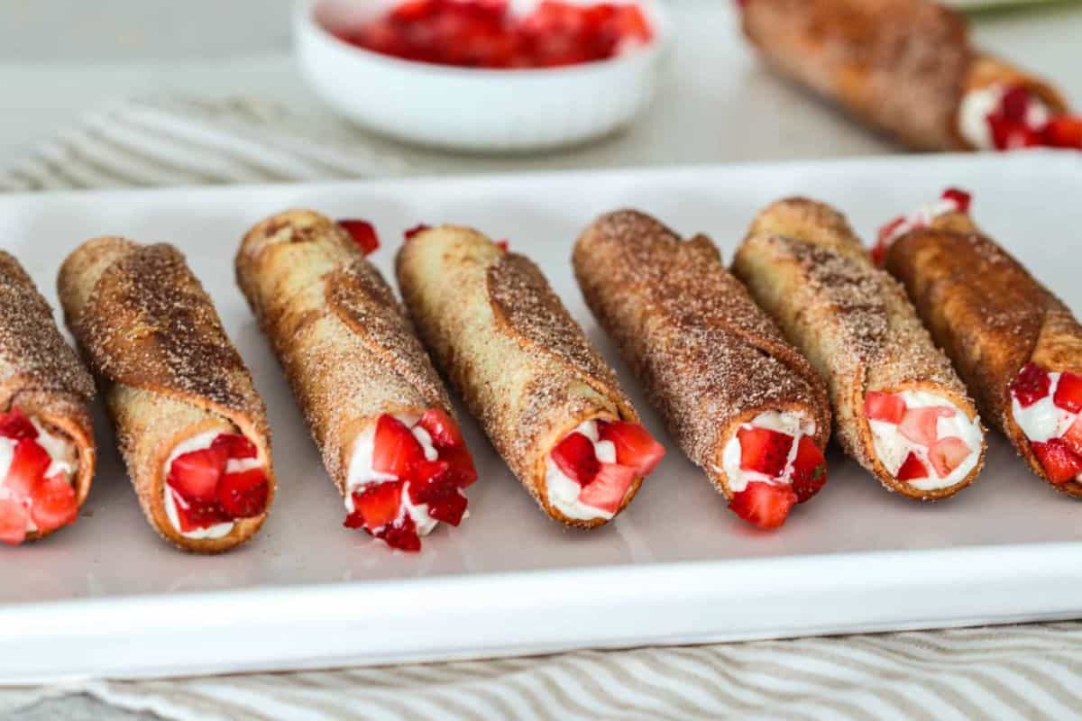 Golden-brown cheesecake cigar rolls filled with sweet cream and diced strawberries, coated in cinnamon sugar and lined up on a white rectangular plate.