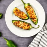 Three air fryer jalapeño poppers with melted cheese sit on a white plate, surrounded by whole and sliced jalapeños, on a gray surface next to a striped cloth.