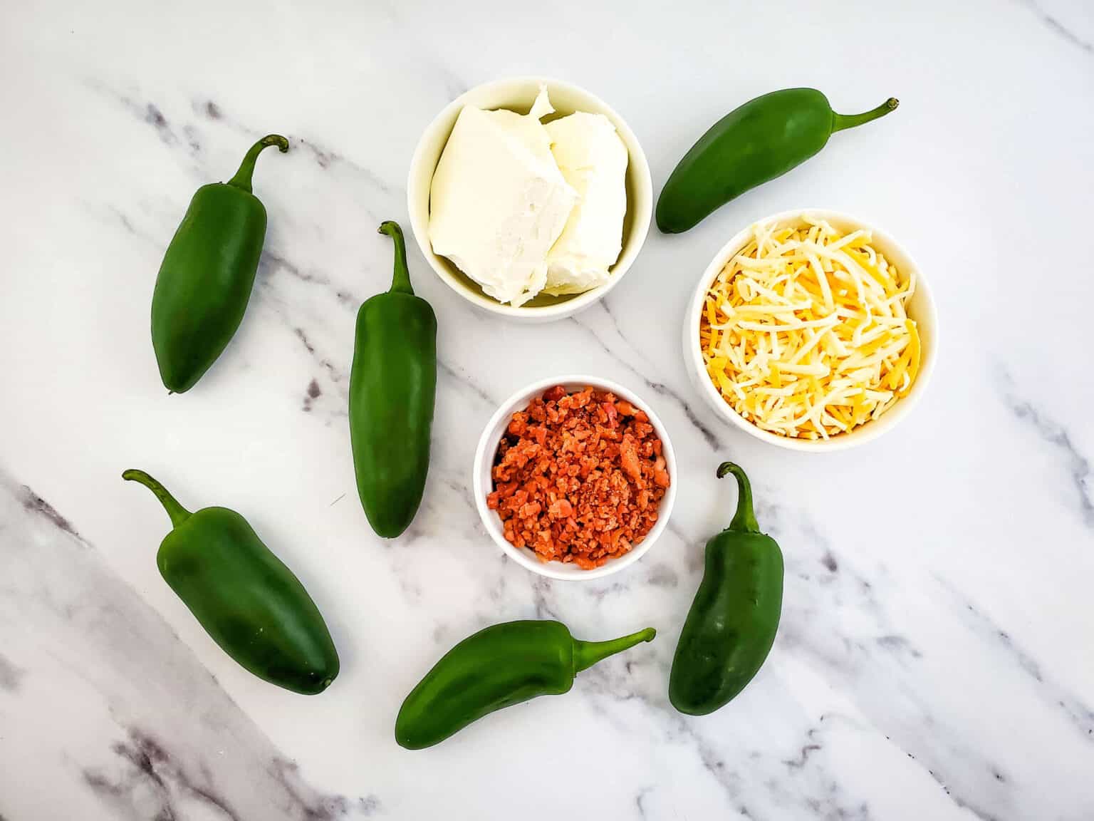 Ingredients for air fryer jalapeno poppers on a marble surface: fresh jalapeños, bowls of cream cheese, shredded cheese, and crumbled bacon.