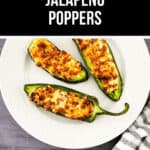 Three air fryer jalapeño poppers with melted cheese rest on a white plate, placed on a gray surface alongside a striped cloth and another jalapeño nearby.