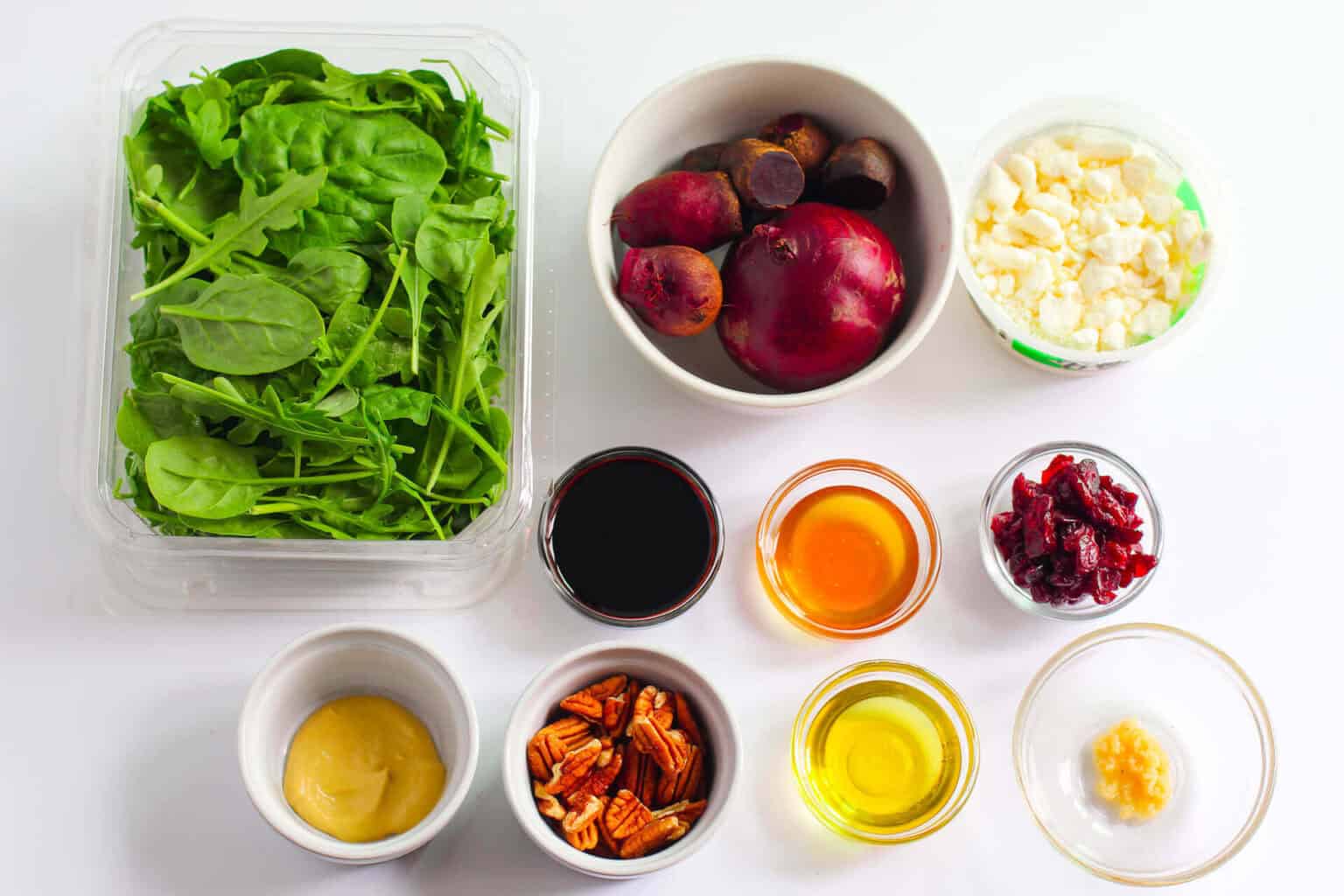 Ingredients  on a white surface, featuring spinach, apples, pecans, cranberries, olive oil, and mustard dressing.