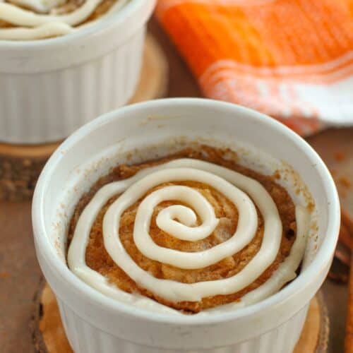 A small ramekin with a baked cinnamon roll, reminiscent of a Cinnamon Mug Cake, topped with a swirl of white icing and placed on a wooden coaster; another ramekin and an orange-striped towel appear in the background.