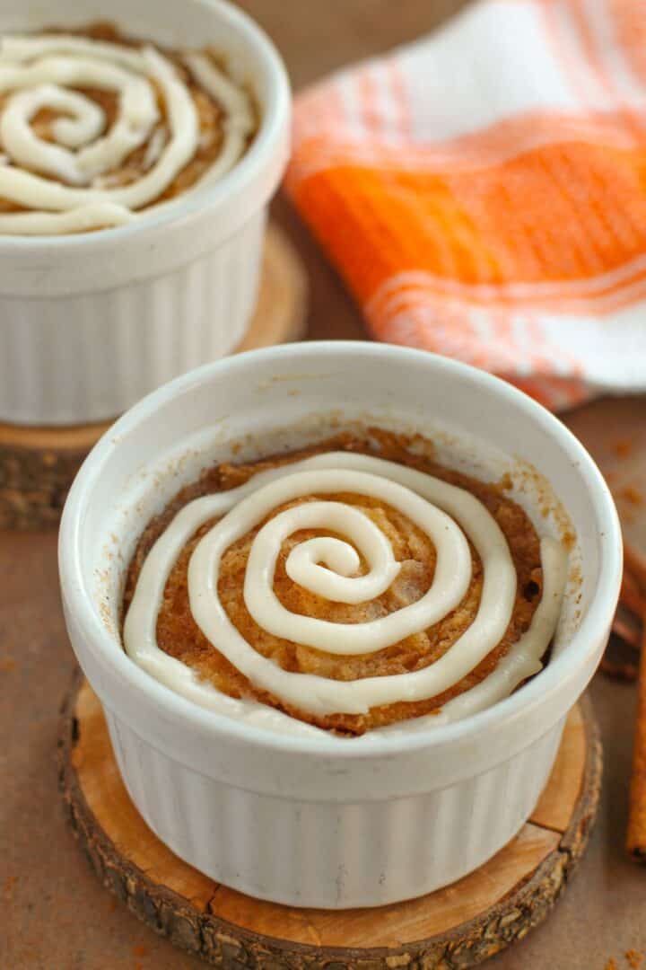 A small ramekin with a baked cinnamon roll, reminiscent of a Cinnamon Mug Cake, topped with a swirl of white icing and placed on a wooden coaster; another ramekin and an orange-striped towel appear in the background.
