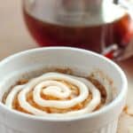 A white ramekin filled with a Cinnamon Mug Cake topped with a swirl of white icing, with a glass container of dark liquid and an orange cloth in the background.