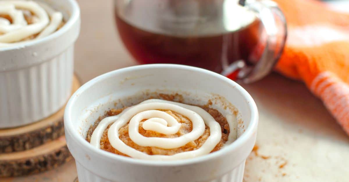 A white ramekin filled with a Cinnamon Mug Cake topped with a swirl of white icing, with a glass container of dark liquid and an orange cloth in the background.