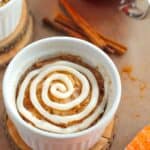 A single-serving dessert in a white ramekin, topped with a spiral of white icing, sits on a wooden coaster. Cinnamon sticks and a cup of tea are in the background.